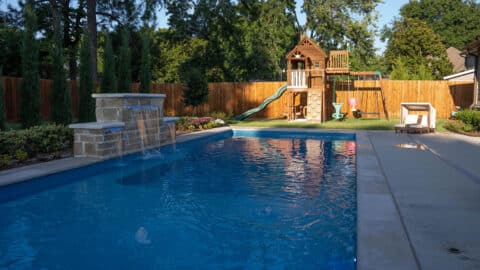 A pool with a waterfall in a backyard with wonderful landscaping and a jungle gym.